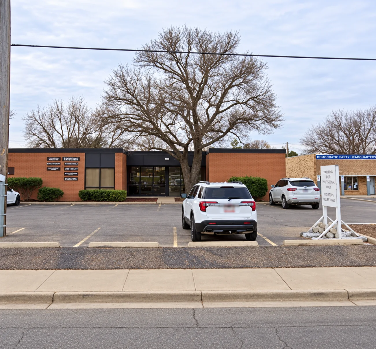 Brick office building viewed across its parking lot, with a central entrance and a large tree